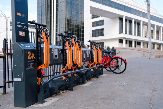 Rostov-on-Don, Russia - October 23, 2019: A row of orange electric scooters and a red bike docked at a rental station with a white modern theater building featuring tall columns in the background