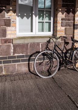 A vintage black bicycle rests against the stone-walled exterior of a building, positioned beneath a window in the warm light of day.