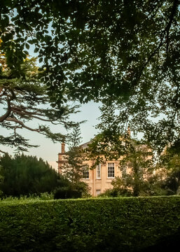 A historic Georgian manor house, captured at golden hour, is framed by the lush foliage of surrounding trees and a manicured hedge.