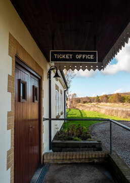 A quiet and serene exterior view of a ticket office at a rural train station, featuring a hanging sign above a wooden door and a peaceful view of the surrounding countryside.