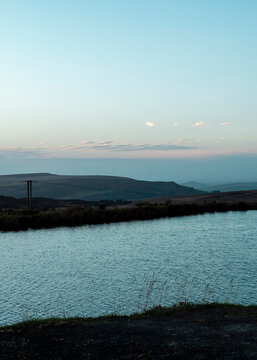 A tranquil scene featuring a calm reservoir or pond in the foreground, set against a vast, rolling moorland landscape during the soft light of twilight, capturing the serene and untouched atmosphere.