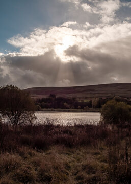 A peaceful and atmospheric landscape view of a calm lake nestled between rolling, grassy hills under a dramatic sky, where brilliant shafts of sunlight break through thick, grey clouds to illuminate.