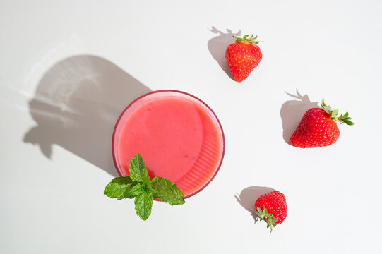 Healthy strawberry smoothie in a glass cup on a white table with morning shadows.