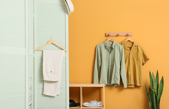 Minimalist home interior with casual mens clothing on hangers and a wooden shoe rack against a yellow wall, a wooden shoe rack holds various footwear, while a straw hat rests on a mint