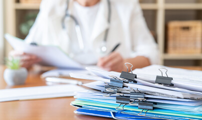 Obraz na płótnie Canvas Healthcare professional wearing a stethoscope is reviewing patient records and administration documents, with a stack of paperwork on the desk showing the volume of work