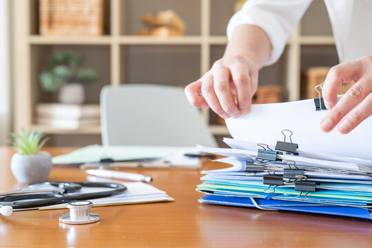 Healthcare personnel managing a stack of overflowing medical records and patient files on a wooden desk, emphasizing administrative tasks and documentation in a healthcare setting