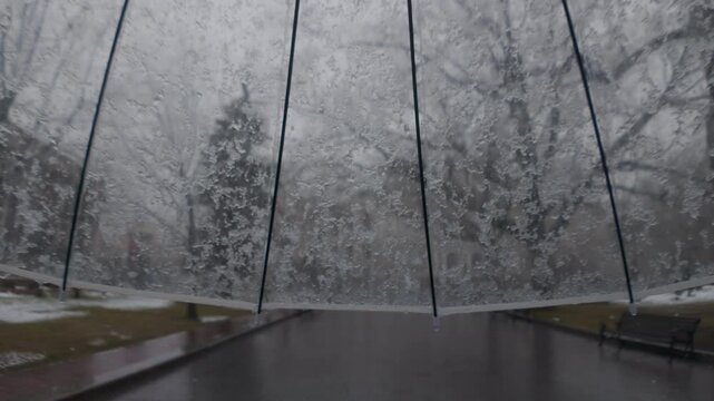 First person view of walking with transparent umbrella under spring sleet falling on gloomy gray day, slow motion