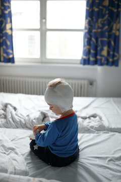 Young child recovering in a pediatric hospital room after cochlear implant surgery. Toddler wearing a protective head bandage while sitting on a hospital bed during postoperative recovery