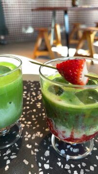 Two glasses of iced matcha latte, one with strawberry, sitting on a table in a modern cafe