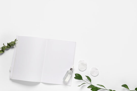 Open blank notebook with eucalyptus and ruscus leaves, cosmetic serum dropper bottle, and cotton pads on white background