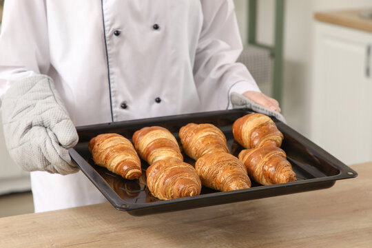 Baker holding baking tray with delicious croissants in kitchen, closeup