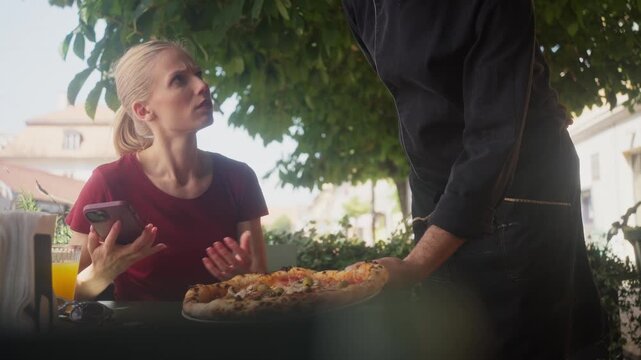 Disappointed blond woman rejecting pizza served by waiter in outdoor restaurant
