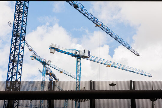 Industrial view of several blue crane structures operating at a building site against a cloudy sky seen through a fence. Stock photo