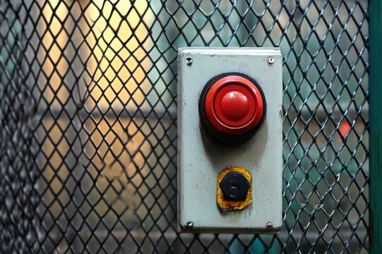 red emergency stop button and small black switch on a weathered metal control box mounted to wire mesh fence in an industrial setting, evoking alertness and caution