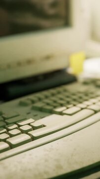 A close view of an old computer keyboard with a monitor in the background. The keyboard shows signs of use. The workspace has a casual setup with some items visible.