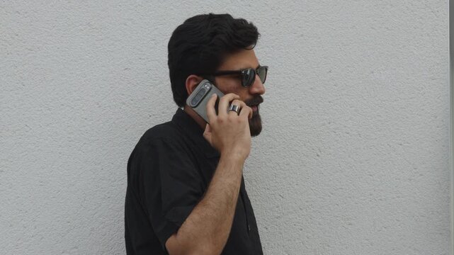 Young bearded man in sunglasses talking on phone by wall