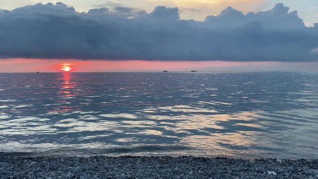 sun sets beneath a dark storm front over a calm sea, casting vivid orange and pink reflections across rippling water while distant boats move slowly along the glowing horizon