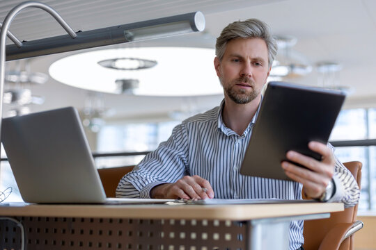 Smiling busy young employee, young entrepreneur using tablet computer, happy businessman executive looking at tab device analyzing finance trading market data working sitting at office desk.