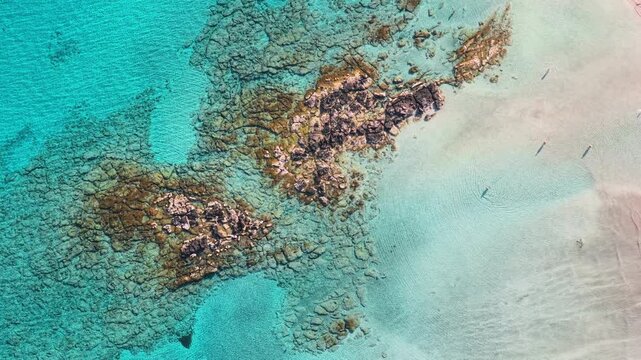 Overhead aerial drone view of a rocky reef in crystal-clear turquoise shallows beside pale sand, Elafonissi, Greece