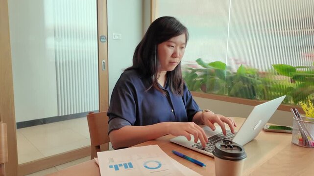 A business professional works on a laptop at a tidy desk while reviewing printed documents in a comfortable office.