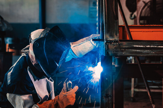 Welder in dark overalls and a full-face mask works intensely. Bright sparks fly from the welding torch as they join metal. Industrial metal structures form the workshop background