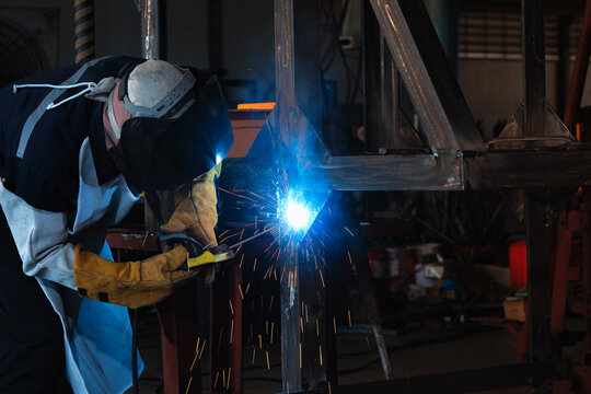 Welder wears a helmet and gloves. Bright blue sparks fly from the welding torch. He focuses on joining metal beams. Industrial factory work. Skilled craftsmanship