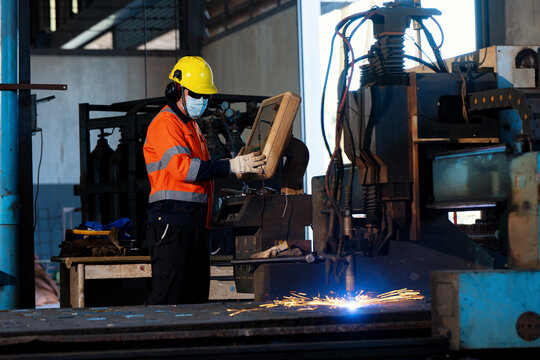 Worker wears a bright yellow hard hat and orange safety vest. They use gloves while handling a metal object near a machine. Sparks fly from the industrial equipment. The person appears focused