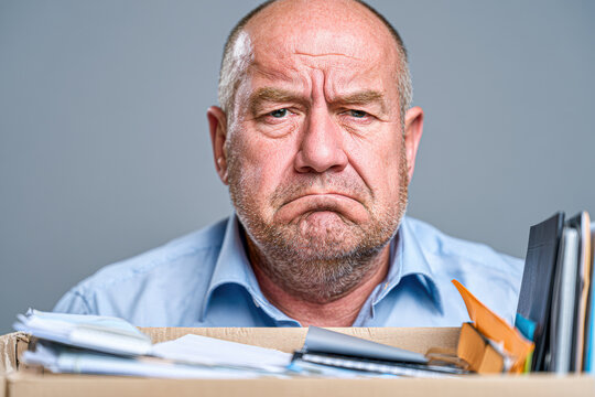 Middle-aged man with a serious expression holding a box of personal belongings, suggesting job loss or resignation in a professional setting.