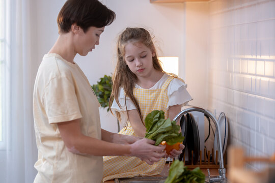 Mother and daughter calmly washing green leafy vegetable in bright modern kitchen, preparing healthy vegetarian food together near window with natural lighting, showing quiet concentration