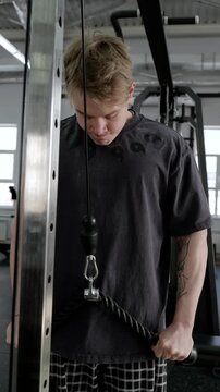 Young athletic man performing rope triceps pushdown on cable machine inside modern gym, focused strength training moment with bent elbows, firm grip, forearm tension, mirrors, and overhead lights