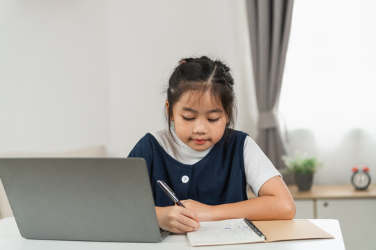 Young Asian girl focused on homework or study project sitting at white desk with laptop and notebook in bright modern room with natural light and home decor