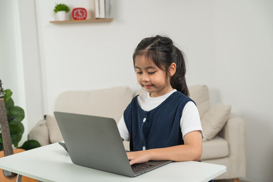 Young girl sitting at a white table using laptop computer focused on screen in modern living room with beige sofa and plant in background for home learning