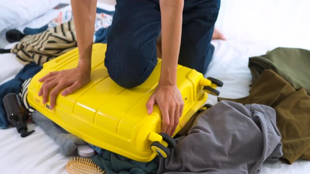 Close-up of Asian woman sitting on an overpacked yellow suitcase to close it. Concept of overpacking issues, travel preparation struggles and holiday planning.
