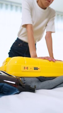 Close-up of Asian woman sitting on an overpacked yellow suitcase to close it. Concept of overpacking issues, travel preparation struggles and holiday planning. Vertical Shot