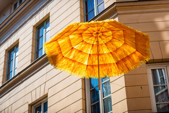 Low-angle close-up of a vibrant yellow and orange raffia fringed beach umbrella suspended from a wire against a dark Norman half-timbered building facade, .
