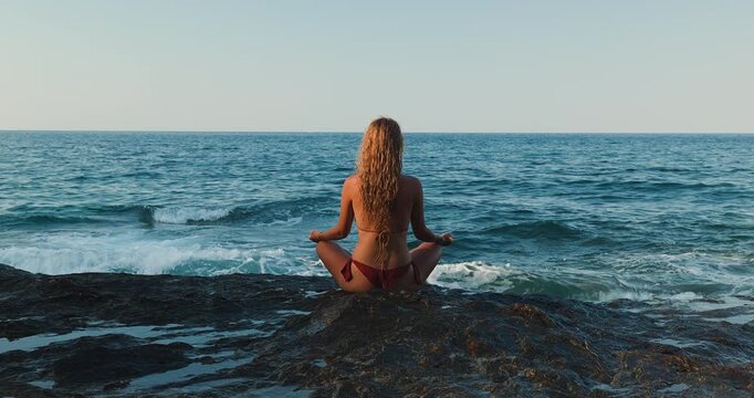 Blonde woman practicing yoga and meditation on coastal rocks by the peaceful ocean waves during a serene summer morning, embracing health, wellness, and mindful relaxation in nature.