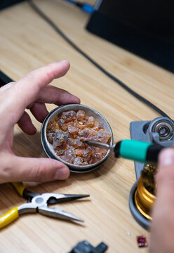 Close-up of a technician's hands holding a metal tin container full of solid pine rosin resin crystals and dipping a hot soldering iron tip into it on a wooden workbench.