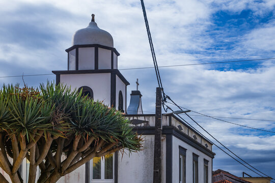 Plaza empedrada con un gran drago frente a una peque&ntilde;a iglesia blanca con bordes oscuros bajo un cielo nublado, t&iacute;pica arquitectura azoriana.