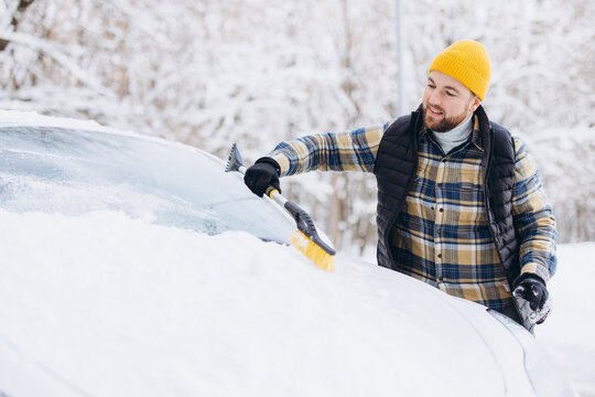 Young man clearing snow and ice from car windshield with a scraper and brush on a cold winter day