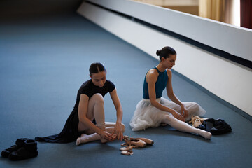 Two ballet dancers preparing pointe shoes and stretching during a quiet rehearsal in a studio hallway © Nikola Spasenoski