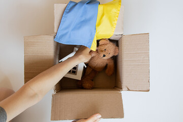 An open cardboard box with a teddy bear, a toy house, and the Ukrainian flag, and a woman's hands on the theme of moving, delivering things, family, hope © Виктория Котлярчук
