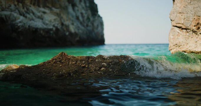 Gentle Ocean Waves Caress a Rugged Rock in Crystal-Clear Turquoise Water, Surrounded by Majestic Cliffs Under a Bright Summer Sky