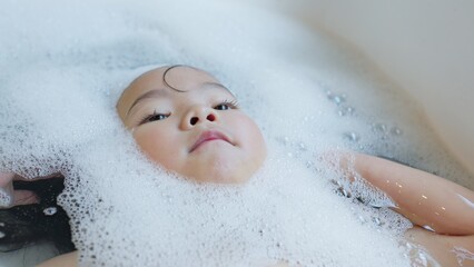 Fototapeta premium A toddler relaxes in a bubble bath, their face emerging from the foam, representing a calm moment of childhood hygiene and sensory wellbeing.