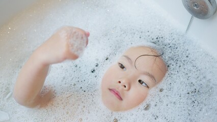 Fototapeta premium A curious young child unwinds in a bathtub full of bubbles, immersed in a peaceful sensory experience as part of a calming daily bathtime routine at home.
