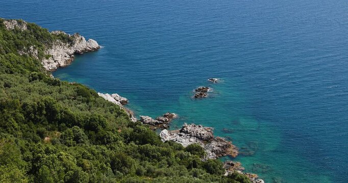 A stunning aerial view of the serene Mediterranean coastline, featuring crystal-clear turquoise waters lapping against rugged rocks and lush green cliffs, bathed in bright sunshine.