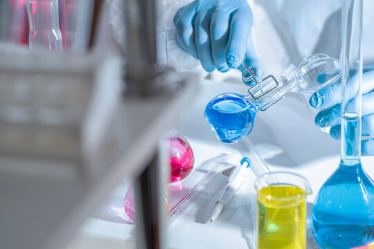 Chemistry laboratory scene with colorful chemical solutions in glass flasks and cylinders, scientist in gloves writing results, analytical experiment workspace.