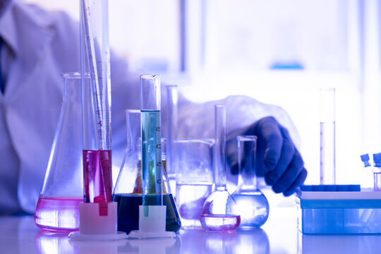 Chemistry laboratory scene with colorful chemical solutions in glass flasks and cylinders, scientist in gloves writing results, analytical experiment workspace.