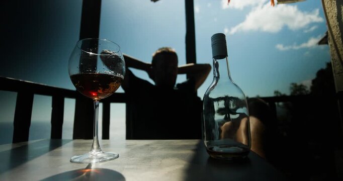 Silhouette of a person relaxing on a sunny balcony with an empty bottle and a glass of drink, enjoying a peaceful moment of solitude and contemplation.