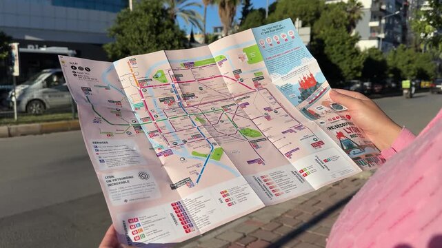 Woman in pink shirt examines city map outdoors, revealing detailed street layout and public transport routes under bright sunlight in urban setting