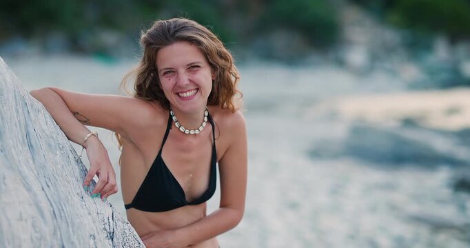 A joyful young woman in a black bikini and shell necklace smiles and laughs happily while leaning on a piece of driftwood at a sunny beach during her summer vacation.